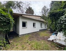 Weiße, einstöckige Gartenlaube mit flachem Satteldach, das teilweise mit Holz verkleidet ist. Die Umgebung ist dicht bewachsen mit Bäumen und Sträuchern, während der Boden teils aus Gras und Erde besteht; im Vordergrund liegen einige Gartenutensilien und Abdeckplanen.