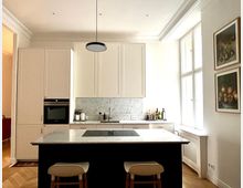 Moderne Küche mit weißem Wandschrank, einer Arbeitsplatte aus Marmor und integrierter Spüle. Im Vordergrund steht eine Kücheninsel mit dunkler Unterseite, heller Marmoroberfläche und zwei Barhockern; an der Wand rechts hängen zwei gerahmte Stillleben, und ein großes Fenster lässt natürliches Licht herein.