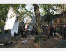Filmset in einem alten Friedhof mit Grabsteinen und Mausoleen im Hintergrund. Beleuchtungsequipment, Kabel und Crewmitglieder sind im Vordergrund zwischen Bäumen und Laub verteilt.