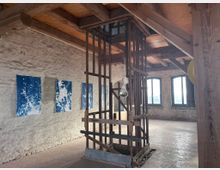 Industrieähnlicher Raum mit sichtbaren Holzbalken und einer rauen, hellen Steinmauer, an der blaue, abstrakte Bilder hängen. In der Mitte des Raums befindet sich eine hölzerne, käfigartige Struktur, und im Hintergrund sind mehrere Fenster mit schwarzem Rahmen zu sehen.