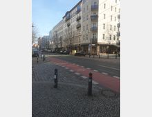 Straßenansicht in einer städtischen Umgebung mit mehrstöckigen Wohngebäuden im klassischen Baustil. Im Vordergrund sind ein mit Kopfstein gepflasterter Gehweg, abgetrennte Fahrradwege sowie eine Straßenbahntrasse zu sehen, eingefasst von Pollern.