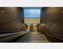 Treppe und Rolltreppe in einem unterirdischen Zugang, umgeben von dunkelbraunen Ziegelsteinwänden. Im oberen Bereich befindet sich ein Fenster aus Glasbausteinen, das diffuses Tageslicht hereinscheint.