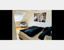 Helles Schlafzimmer mit Dachschrägen, Holzboden und einem großen Bett mit schwarzer Bettwäsche. An der Wand hängt ein Schwarz-Weiß-Bild, und ein Sessel sowie ein kleines Tischchen stehen unter einem Dachfenster mit Blick nach draußen.