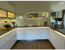 Eine moderne Küche mit weißen, schlichten Fronten und einer dunklen Arbeitsplatte. Eine indirekte Beleuchtung unter den Schränken und Regalen sorgt für Akzente; der Raum enthält Deko-Elemente wie Pflanzen, ein Messerblock, und eine Küchenmaschine auf der Arbeitsfläche.