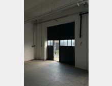 Innenraum einer Industriehalle mit weiß gestrichenen Backsteinwänden und Betonboden; an der Decke verlaufen sichtbare Rohre und eine lange Leuchtstofflampe. Eine große schwarze Schiebetoröffnung mit Fenstersegmenten lässt Tageslicht herein und gibt den Blick nach draußen auf einen eingezäunten Bereich frei.