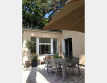 Heller Außenbereich eines Hauses mit beigem Putz, großen Fenstern und einer Terrasse mit heller Steinpflasterung. Auf der Terrasse stehen ein grauer Tisch mit mehreren passenden Stühlen und Topfpflanzen; darüber ein großer Sonnenschirm, im Hintergrund Bäume.