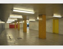 Unterirdischer Gang mit gefliesten Wänden in Weiß und senfgelben Mosaiksäulen. Der Boden besteht aus hellen Fliesen, die Decke ist niedrig mit rechteckigen Leuchtstofflampen, und am Ende sind eine rote Wand und eine gelbe Tür sichtbar.