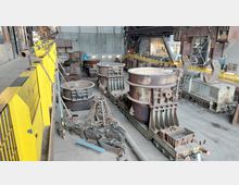 Industriehalle mit rostigen Stahlbehältern und Maschinen auf Schienen, umgeben von gelben Schutzgeländern. Im Hintergrund sind metallische Strukturen, ein Kranhaken und eine kleine Lokomotive sichtbar.