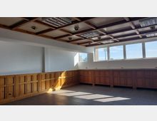 Großer, leerer Raum mit Holzvertäfelung an den Wänden, einer Decke in Kassettenoptik mit eingebauten Leuchten und rechteckigen Fenstern, die Tageslicht hereinlassen. Der Boden besteht aus dunklen Fliesen, die kompakte, schlichte Gestaltung prägen.