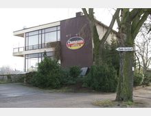 Zweistöckiges Gebäude mit großen Fenstern und brauner Fassade, mit einem Schild "Champions Teehaus" an der Wand. Im Vordergrund befinden sich Bäume, Sträucher und ein Wegweiser mit der Aufschrift "Zum Geläuf".