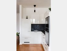 Moderne Küche mit weißen Hochglanzschränken, einer dunklen Arbeitsplatte und einer schwarzen Rückwand. Im Vordergrund befindet sich ein Spülbecken, rechts ein eingebauter Ofen, und oben hängt eine schwarze Pendelleuchte.