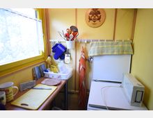 Kleine Küche mit gelb gestrichenen Wänden, gelbem Fensterrahmen und einer Gardine. Auf einem schmalen Holzarbeitsplatz stehen Küchenutensilien, ein Schneidebrett und Tassen; an einer Hakenleiste hängen Kochhandschuhe, Besteck und ein Handtuch. Rechts steht ein kleiner Kühlschrank mit einer karierten Abdeckung.