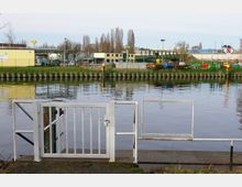 Blick auf einen Fluss mit einer weißen Metallabsperrung im Vordergrund. Im Hintergrund befinden sich industrielle Gebäude, Bauzäune, Bäume sowie Arbeitsgeräte auf einem eingezäunten Gelände.