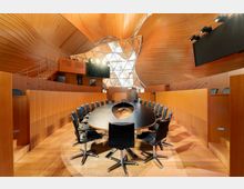 Moderner Konferenzraum mit einem ovalen Tisch, umgeben von schwarzen Lederstühlen auf Rollen. Die Wände und Decke bestehen aus geschwungenem, warmem Holz, während ein großflächiges Fenster mit geometrischen Mustern Tageslicht hereinlässt; Mikrofone und Bildschirme sind installiert.