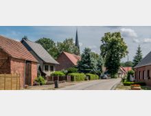 Ländliche Straße mit Backsteinhäusern und gepflegten Vorgärten, gesäumt von Bäumen und Sträuchern; im Hintergrund ist der Turm einer Kirche mit spitzer Dachform sichtbar.