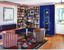 Wohnzimmer mit Holzfußboden, einer blauen Bücherwand und zwei gestreiften Sesseln. Im Raum befinden sich ein niedriger Tisch mit Blumenvasen, ein bunter Teppich und zwei Fenster, die Tageslicht hereinlassen.