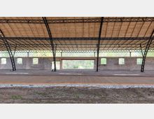 Offene Industriehalle mit einem Dach aus Holzpaneelen, gestützt von schwarzen Stahlträgern. Die Backsteinwände mit mehreren Fensteröffnungen und ein großer rechteckiger Durchgang geben den Blick auf eine bewaldete Umgebung frei.