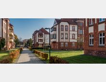 Mehrstöckige Wohngebäude aus rotem Klinker mit weißen Akzenten und Satteldächern, die um einen gepflasterten Weg mit kleinen Hecken und Rasenflächen angeordnet sind. Im Hintergrund befinden sich Bäume und parkende Autos.