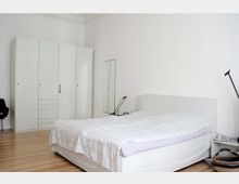 Schlafzimmer mit weiß gestrichenen Wänden, einem großen weißen Kleiderschrank, einem Doppelbett mit weißem Bettbezug und einem Nachttisch. Der Boden besteht aus hellem Holz, und an der Wand hängt ein schmaler Spiegel.