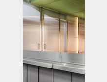 Eine Innenansicht mit glatten, metallischen Oberflächen: Im Vordergrund eine graue Metalltheke, dahinter halbtransparente Wände mit goldfarbenen Rahmen und Griffen. Die Decke ist mit einem grünlichen, strukturierten Gittermuster versehen.