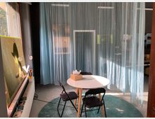 Ein kleiner, heller Raum mit einem runden weißen Tisch und zwei schwarzen Stühlen, umgeben von halbtransparenten blauen Vorhängen. Auf der linken Seite stehen Regale und ein Fenster, das Licht in den Raum lässt.