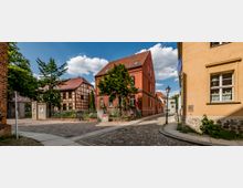 Straßenszene in einer historischen Altstadt mit Kopfsteinpflaster, roten und gelben Backsteinhäusern sowie einem Fachwerkhaus im Hintergrund. Ein kleiner Baum und Zaunelemente rahmen den Vorgarten eines Gebäudes, während eine Laterne und schmale Gehwege die Szene ergänzen.