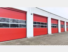 Außenansicht eines Gebäudes mit mehreren roten Garagentoren, die mit mittig verlaufenden Fenstereinsätzen versehen sind. Der Boden besteht aus einem gepflasterten Vorplatz, und die Wände des Gebäudes sind weiß.