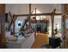 Großzügiger Wohnraum mit sichtbaren Holzbalken und Dachschrägen, einer offenen Küche mit zentraler Kücheninsel, Holzfußboden und Galeriebereich. Im Vordergrund steht ein Kaminofen, rechts ist ein Stapel Holz gelagert, und links befinden sich Dachfenster, die den Raum mit Tageslicht durchfluten.