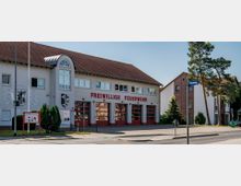 Zweistöckiges Gebäude einer freiwilligen Feuerwehr mit rotem Ziegeldach und mehreren großen Garagentoren, auf deren Fassade „Freiwillige Feuerwehr“ steht. Davor befinden sich ein Gehweg, ein Straßenschild mit der Aufschrift „Hirschweg“ sowie einige Bäume und Sträucher.