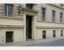 Fassade eines Altbaus mit gelb-beigem Putz, mehreren rechteckigen Fenstern und einem eleganten Eingangsportal mit teilweise geöffneter Holztür. Im unteren Bereich sind einige Graffiti auf Wänden sowie vergitterte Kellerfenster sichtbar; rechts davor verläuft ein Gehweg mit Bordstein.