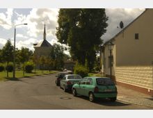 Straßenszene in einer ländlichen Gegend mit geparkten Autos entlang einer asphaltierten Straße; im Hintergrund eine kleine Kirche mit achteckigem Turm und Spitzdach, umgeben von Grünflächen und Bäumen. Rechts im Bild ist ein Haus mit cremefarbener Fassade und kleinen Fenstern zu sehen.