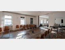 Ein einfacher Raum mit weißen Wänden, Holzdielenboden und mehreren Holzstühlen in Reihen vor Fenstern mit weißem Rahmen. Im Hintergrund stehen technische Geräte, ein Tisch mit Computerausrüstung und Stative, die auf eine Nutzung als Veranstaltungs- oder Probenraum hinweisen.