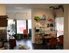 Ein gemütlicher Innenraum mit Café-Atmosphäre, ausgestattet mit bunten Möbeln, Regalen voller Souvenirs und Getränken sowie einer Dekoration mit Pflanzen und verspielten Elementen. Im Hintergrund ist eine gläserne Eingangstür mit dem Schriftzug "BAR" zu sehen, daneben stehen ein kleines Tisch- und Stuhl-Set sowie grüne Topfpflanzen.