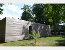 Ein rechteckiges Gebäude mit einer Fassade aus horizontalen Holzpaneelen, umgeben von einer grünen Wiese und dichten Bäumen. Im Vordergrund steht ein kleiner Baum vor einer schmalen, vertikalen Türöffnung.