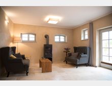 Wohnzimmer mit hellbeigen Wänden und Fliesenboden. Zwei graue Sessel stehen sich gegenüber, mit einem Holz-Couchtisch in der Mitte, einem Kaminofen an der Wand und kleinen Fenstern mit Rundbögen; rechts eine Glastür mit Vorhängen.