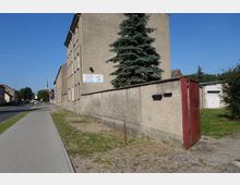 Straßenszene mit breitem gepflastertem Gehweg und mehrstöckigem Wohngebäude aus schlichten Putzfassaden auf der rechten Seite. Im Vordergrund eine gemauerte Mauer mit rotem Metalltor und einer großen Tanne, im Hintergrund ein Kirchturm sowie weitere Gebäude entlang der Straße.