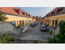 Schmale, gepflasterte Gasse mit gelben Reihenhäusern auf beiden Seiten, roten Ziegeldächern und Fensterläden; Treppen und kleine Höfe führen zu den Eingängen der Häuser, einige Autos parken entlang der Straße. Im Hintergrund sind weitere Gebäude und Bäume sichtbar.