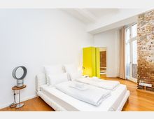 Helles, minimalistisches Schlafzimmer mit Doppelbett und weißer Bettwäsche auf Holzboden, neben einem kleinen Beistelltisch mit Ventilator. Im Hintergrund stehen ein auffällig gelber Kleiderschrank und hohe Fenster mit Vorhängen neben einer freigelegten Ziegelwand.