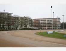 Eine biegsame Sandbahn, vermutlich eine Lauf- oder Pferderennbahn, mit angrenzendem Grünstreifen. Im Hintergrund stehen mehrstöckige Wohngebäude mit Balkonen, Bäumen und Beleuchtungsmasten.