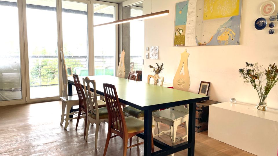Esszimmer mit großem rechteckigem Tisch und verschiedenen Stühlen, modernem Holzboden und einer Wand mit Kunstwerken und dekorativen Elementen. Große Fensterfront mit Blick ins Freie lässt viel Tageslicht herein.