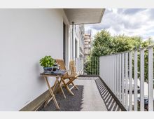 Ein Balkon mit weißer Hauswand und Metallgeländer, ausgestattet mit einem kleinen Holztisch und zwei Holzklappstühlen, auf dem Tisch steht ein Basilikumtopf sowie eine Kaffeekanne. Im Hintergrund sind Bäume, geparkte Autos und ein Gerüst an einem Nachbargebäude zu sehen.