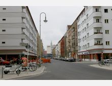 Straßenszene in einer Stadt mit mehrstöckigen Wohngebäuden auf beiden Seiten, teils glatt verputzt, teils mit verzierter Fassade. Im Vordergrund stehen Fahrräder, ein Roller und ein Traktor, während am Straßenrand geparkte Autos und Baustellenabsperrungen sichtbar sind; im Hintergrund ein moderneres Gebäude mit Turm.