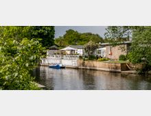Wohngebiet an einem ruhigen Flussufer mit mehreren kleinen Häusern, umgeben von Gärten und Bäumen. Im Vordergrund ist ein blaues Tretboot am Ufer befestigt, und im Hintergrund stehen weiße Zäune und Terrassenmöbel.