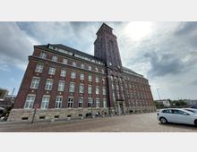 Backsteingebäude mit Turm und historischer Architektur, geprägt durch zahlreiche Fensterreihen, Natursteinsockel und ein mit einer Uhr versehenes Turmgesicht; davor ein gepflasterter Platz mit geparkten Autos.