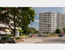Wohnsiedlung mit mehreren mehrstöckigen Wohngebäuden im Hintergrund, darunter ein großes Hochhaus mit Balkonen. Im Vordergrund sind ein Gehweg, parkende Autos, Bäume und einzelne Grünflächen zu sehen.