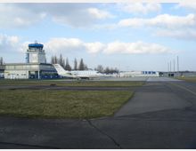 Kleiner Flughafenterminal mit einem mehrstöckigen Kontrollturm und einem geparkten, weißen Privatflugzeug auf dem Rollfeld. Im Hintergrund sind Hangars, flache Gebäude und eine freie Landschaft zu sehen.