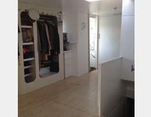 Minimalistisch eingerichteter Innenraum eines kleinen Wohnbereichs mit weißem Interieur, offenem Kleiderschrank mit Regalen und einer Tür, die zu einem hellen Flur führt. Der Boden besteht aus hellem Holz, und Wände sowie Möbel sind schlicht gestaltet.