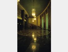 Großer, prunkvoller Innenraum mit marmornem Boden, einem kreisförmigen Muster in der Mitte und herabhängenden Kristallleuchtern. Eine geschwungene Treppe führt nach oben, während hohe Fenster und dekorative Wandpaneele den Raum säumen.