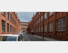 Eine schmale Straße mit parkenden Autos, flankiert von hohen, roten Backsteingebäuden mit großen, weißen Fenstern im Industrial-Stil. Ein Verbindungsbauwerk aus Backstein überspannt die Straße, während Laternen entlang des Gehweges stehen.