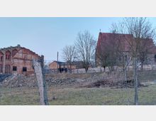 Ländliche Szene mit einer alten Backsteinruine auf der linken Seite, einer kleinen historischen Kirche aus Naturstein mit rotem Ziegeldach rechts und einem eingezäunten Feld im Vordergrund. Im Hintergrund sind weitere kleine Gebäude sichtbar, umgeben von Bäumen.